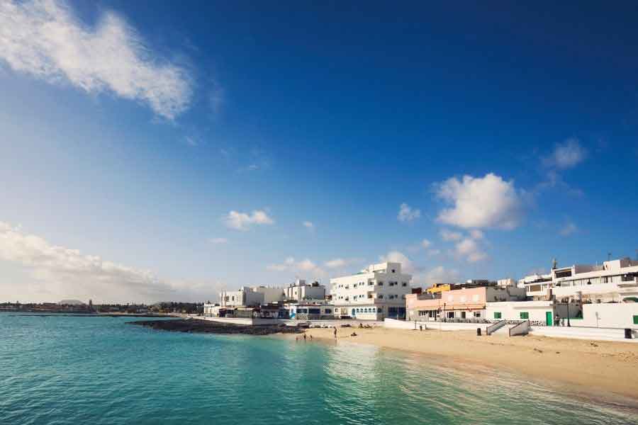 Corralejo, Fuerteventura A quiet beach in Corralejo, Fuerteventura, with turquoise water gently lapping the shore and low-rise whitewashed buildings lining the seafront. A few people relax on the sand under clear skies—an ideal setting for chasing Europe’s winter sunshine.