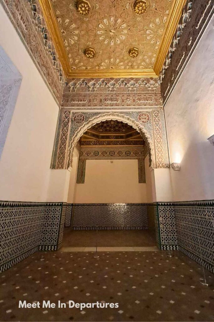 Interior of the Cuarto del Príncipe inside the Real Alcázar of Seville, featuring intricate Moorish tilework on the lower walls, a decorative horseshoe arch with detailed stucco patterns, and a golden coffered ceiling with ornate geometric designs.