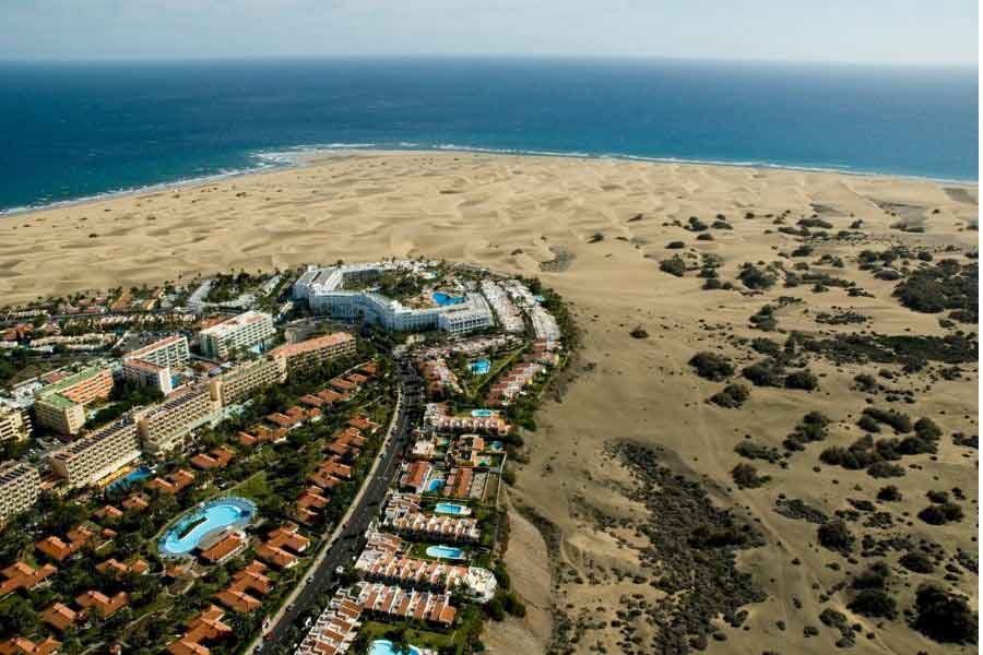 Dunes at Maspalomas Aerial view of Maspalomas in Gran Canaria, showing a striking contrast between the golden sand dunes and the nearby resort buildings with pools. The Atlantic Ocean stretches out in the background, capturing the appeal of southern Europe’s sunny winter escapes.