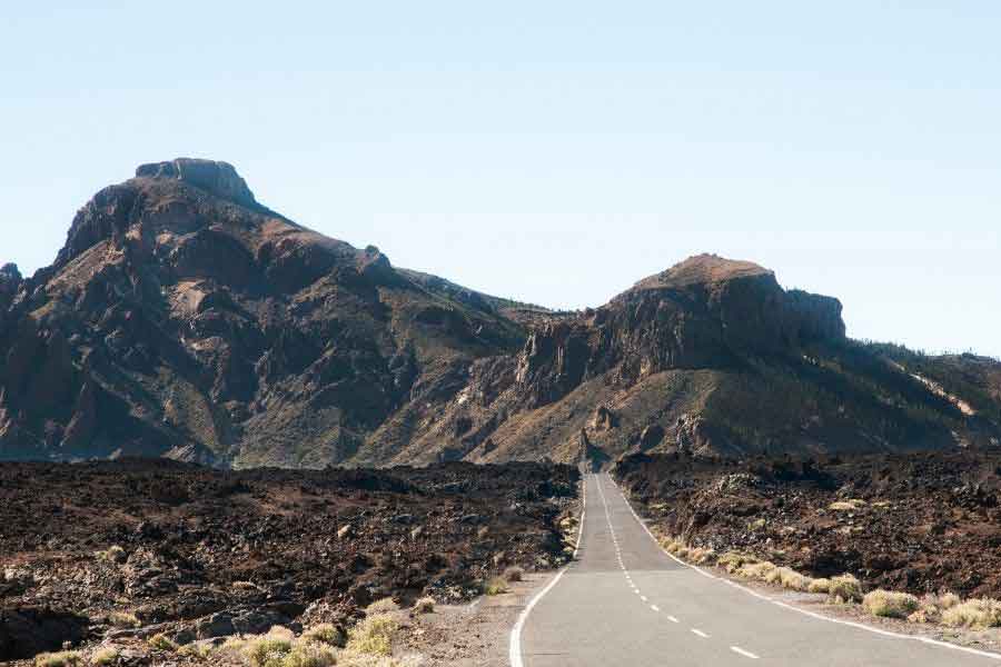 Timanfaya National Park, Lanzarote A long, empty road cutting through the rugged volcanic landscape of Timanfaya National Park in Lanzarote, with dramatic rocky mountains in the distance under a clear blue sky. The stark terrain and warm light capture the otherworldly beauty of winter warmth in southern Europe