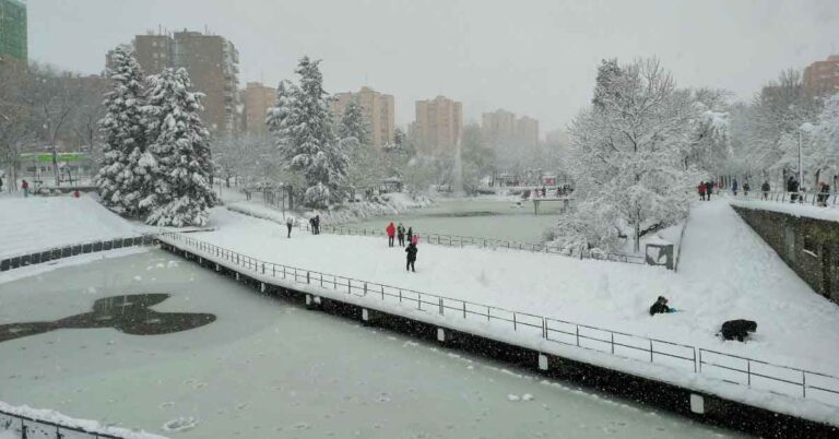 A snowy winter scene in a Madrid city park, where people walk along snow-covered paths beside a frozen pond. Trees are thick with snow, and apartment blocks stand in the background under a grey sky. This rare snowfall visually answers the question, "does it snow in Spain," showing that even cities like Madrid can experience heavy snow in winter.