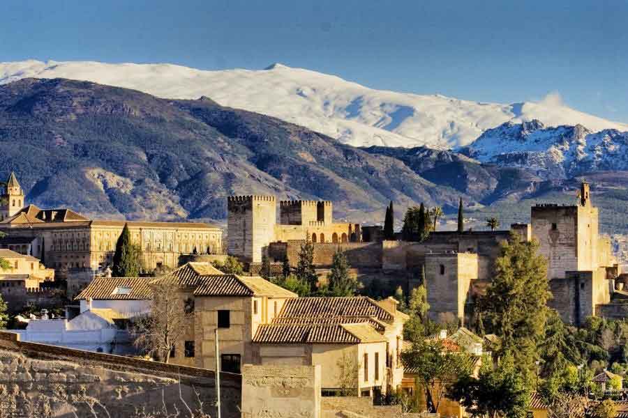 A panoramic view of the Alhambra palace complex in Granada, Spain, with terracotta-roofed houses in the foreground. Behind the historic buildings, the snow-covered Sierra Nevada mountains rise under a clear blue sky. This striking contrast of sunlit architecture and snowy peaks illustrates that yes, it does snow in Spain—especially in regions like Andalusia’s high mountains.