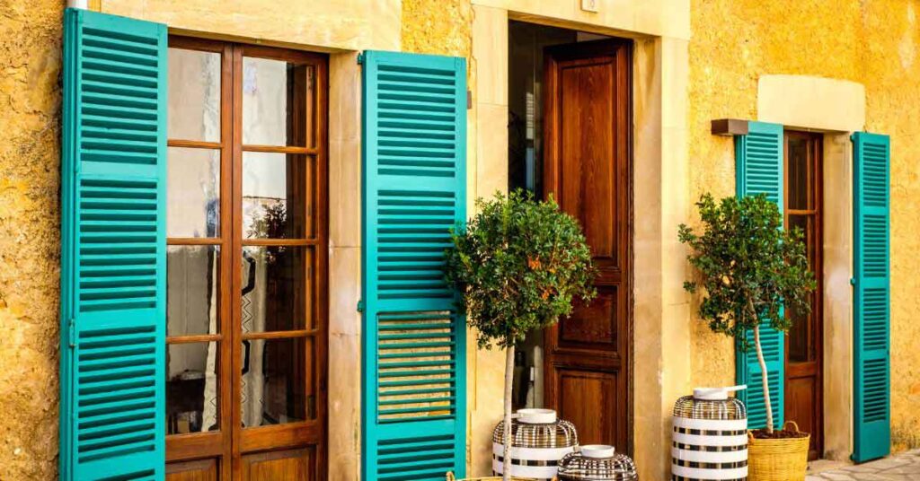 Streets in Mallorca A rustic Mediterranean-style house with bright turquoise shutters, wooden doors, and potted plants in the sun. The warm stone walls and soft lighting create a relaxed atmosphere, evoking the feeling of winter sun in Europe.