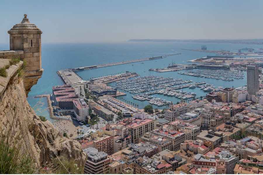 View from Castillo de Santa Bárbara overlooking Alicante city, marina, and Mediterranean coastline on a clear sunny day.