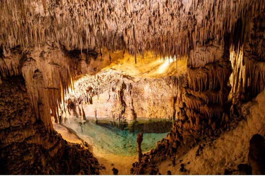 Interior of the Cuevas del Drach in Mallorca, showing dramatic stalactites hanging over a clear underground lake illuminated by warm lighting.