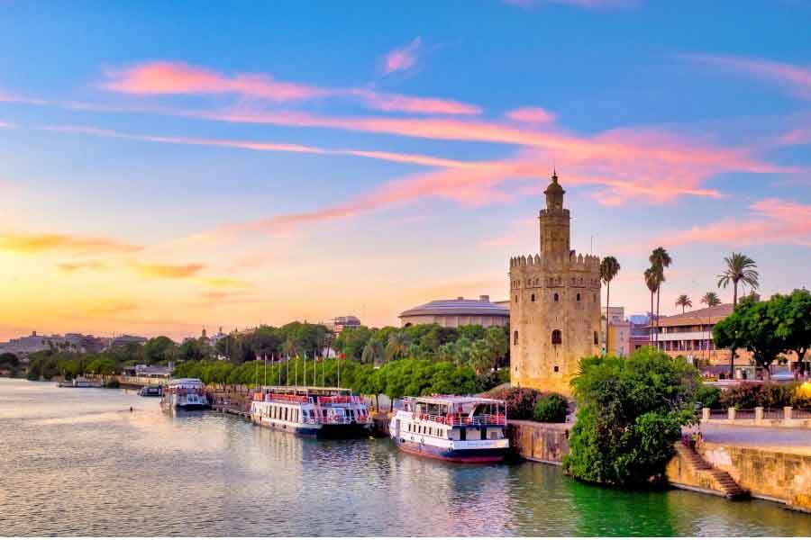 Torre del Oro beside the Guadalquivir River in Seville at sunset, with colourful sky, palm trees, and riverboats moored along the waterfront.