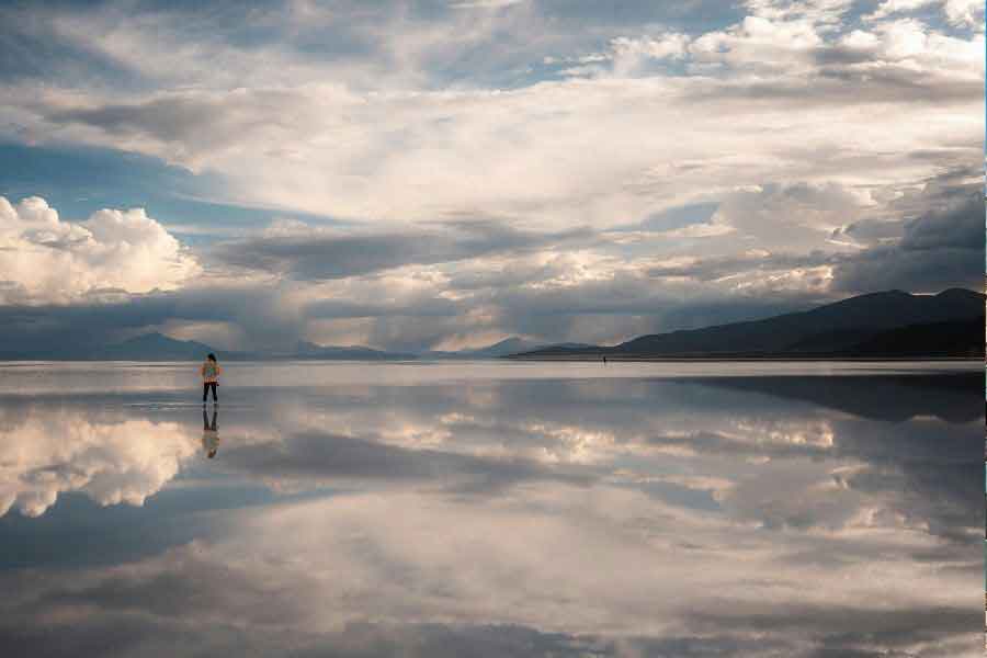 Person standing on the reflective Uyuni Salt Flats in Bolivia, surrounded by dramatic clouds and distant mountains mirrored perfectly on the reflective surface.