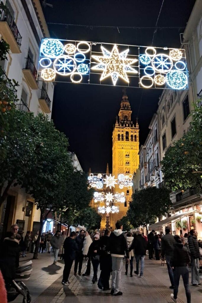 Christmas lights in Seville illuminating the street leading to the Giralda Tower, with people strolling beneath festive decorations and enjoying the evening atmosphere in the city centre.