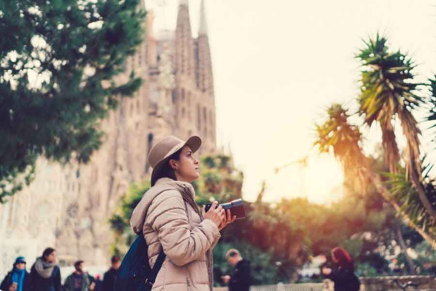 Traveller in Barcelona during winter wearing a padded jacket and hat, holding a camera near the Sagrada Família as the soft afternoon sun highlights Spain’s mild coastal winter weather.