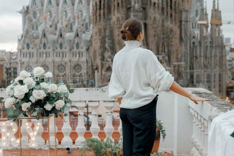 Woman wearing a cosy white jumper and dark trousers on a rooftop terrace overlooking the Sagrada Família in Barcelona, showing typical winter fashion in Spain’s mild Mediterranean climate.