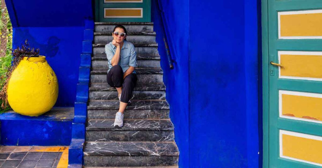 The complete Morocco winter packing list A woman sitting on stone steps at Jardin Majorelle in Marrakech wearing sunglasses, a light denim shirt and cropped trousers — an example of what to wear in Morocco in winter, showing a practical and comfortable outfit for cooler days.