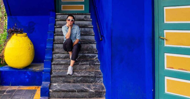 A woman sitting on stone steps at Jardin Majorelle in Marrakech wearing sunglasses, a light denim shirt and cropped trousers — an example of what to wear in Morocco in winter, showing a practical and comfortable outfit for cooler days.