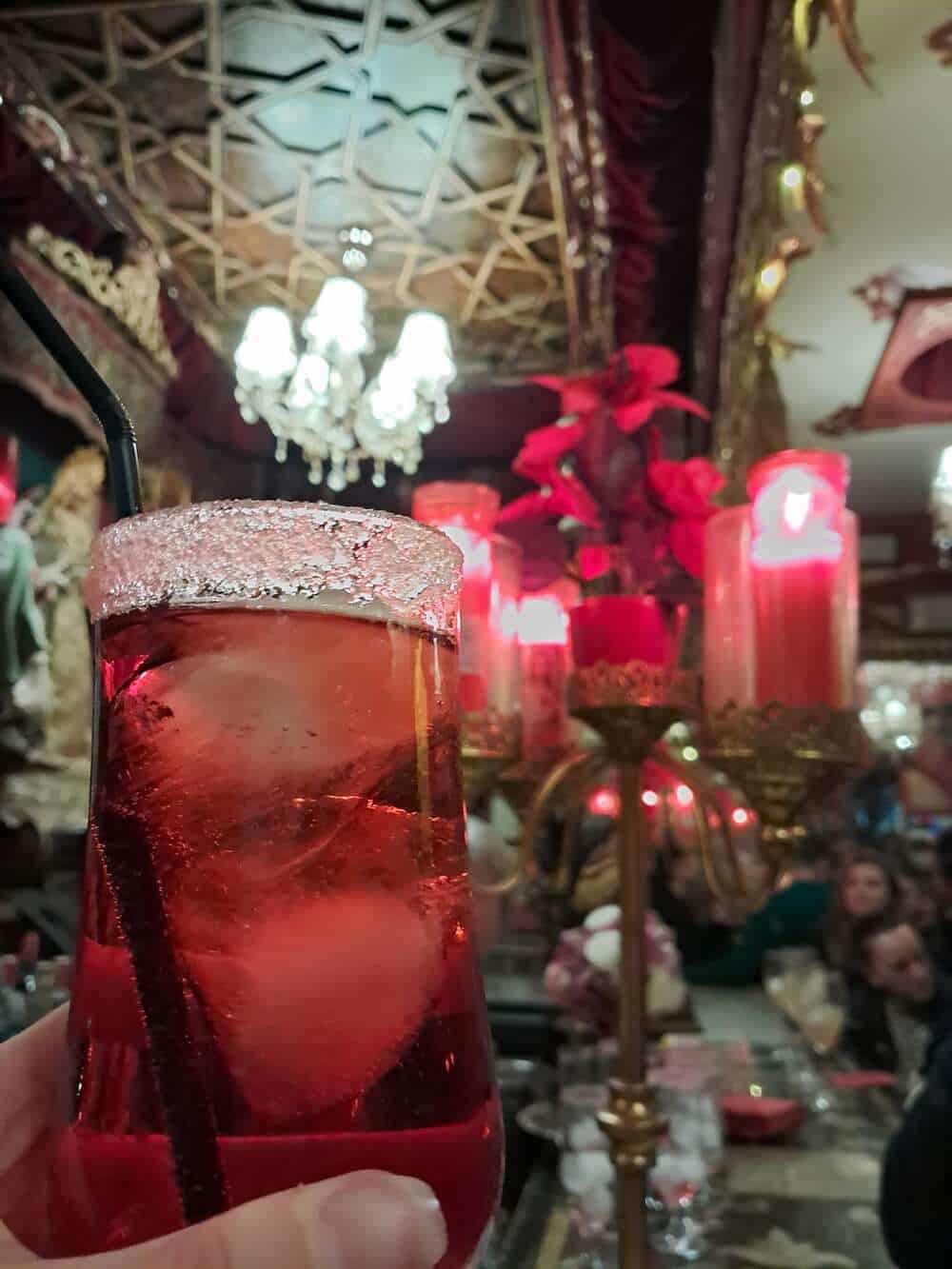 A close-up of the 'Sangre de Cristo' (Blood of Christ) cocktail at Bar Garlochi, a famous kitsch-themed bar in Seville. The red drink is served in a glass with a sugar-rimmed edge and a black straw, set against a backdrop of glowing red candles and ornate religious decor, representing one of the most eccentric things to experience in Seville.