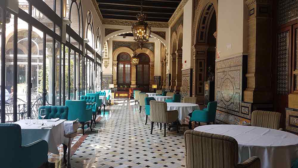The opulent dining room of the Hotel Alfonso XIII in Seville, featuring grand arches, intricate Mudéjar-style wall tiles, and polished marble floors. Teal velvet armchairs and linen-topped tables are arranged along a glass-walled corridor overlooking a courtyard, showcasing one of the most luxurious places to explore in Seville.