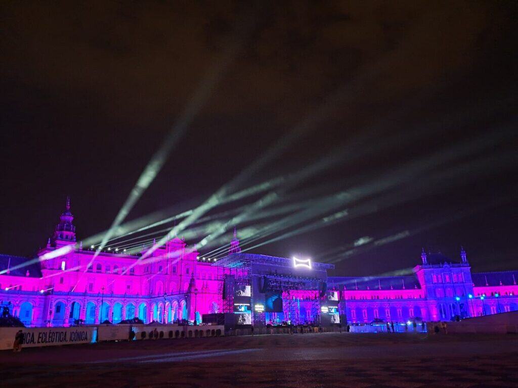The Icónica Sevilla Fest at the Plaza de España at night, featuring the historic neo-Moorish pavilion illuminated in brilliant pink and purple lights. Beams of white spotlights stretch across the dark sky from a large concert stage, showcasing one of the most spectacular outdoor events in Seville.