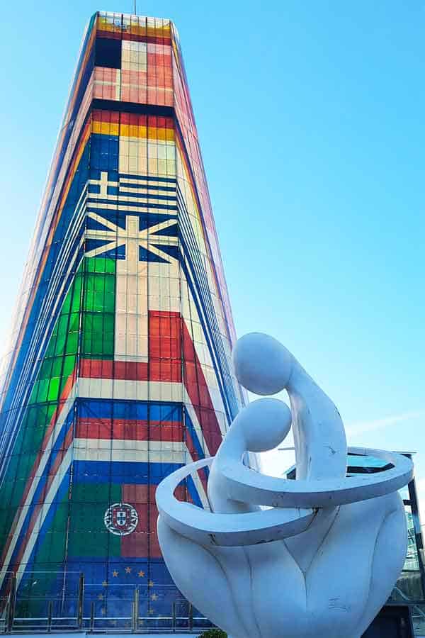 The striking Pabellón de Francia on Isla de la Cartuja, featuring a massive tapered tower decorated with colourful international flags and the European Union emblem. In the foreground, a modern white abstract sculpture of two embracing figures sits under a clear blue sky, representing the more contemporary Seville tourist attractions found outside the historic centre.