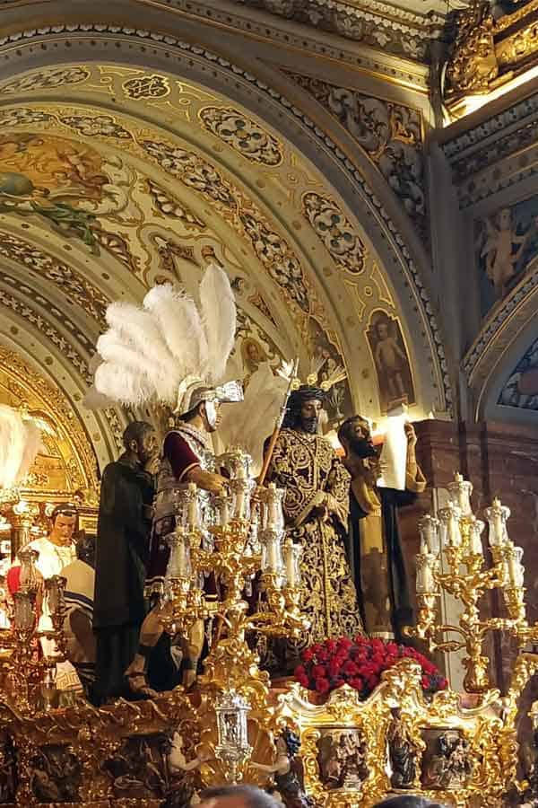 Inside a grand Seville church, an ornate golden "paso" (float) for the Semana Santa processions features lifelike religious statues, including a Roman soldier with white plumes and a figure in embroidered purple robes. The float is adorned with red roses and elaborate candelabras, representing the most deeply-rooted historical traditions in Seville.
