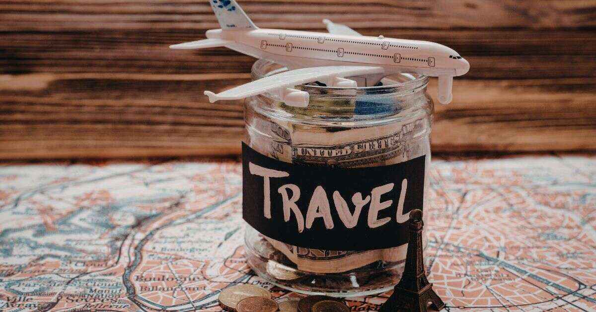 Trip budget calculator image showing a travel savings jar filled with cash and coins, topped with a toy aeroplane, sitting on a map to represent travel planning, holiday costs, trip expenses, and saving money for a trip.
