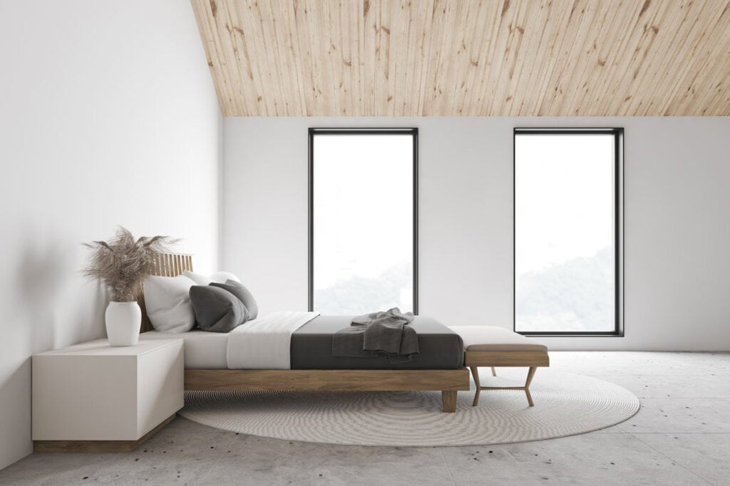 Photo d'une chambre vue de côté. Un lit, une table de chevet, et face à l'objectif deux fenêtres donnant sur un extérieur arboré.