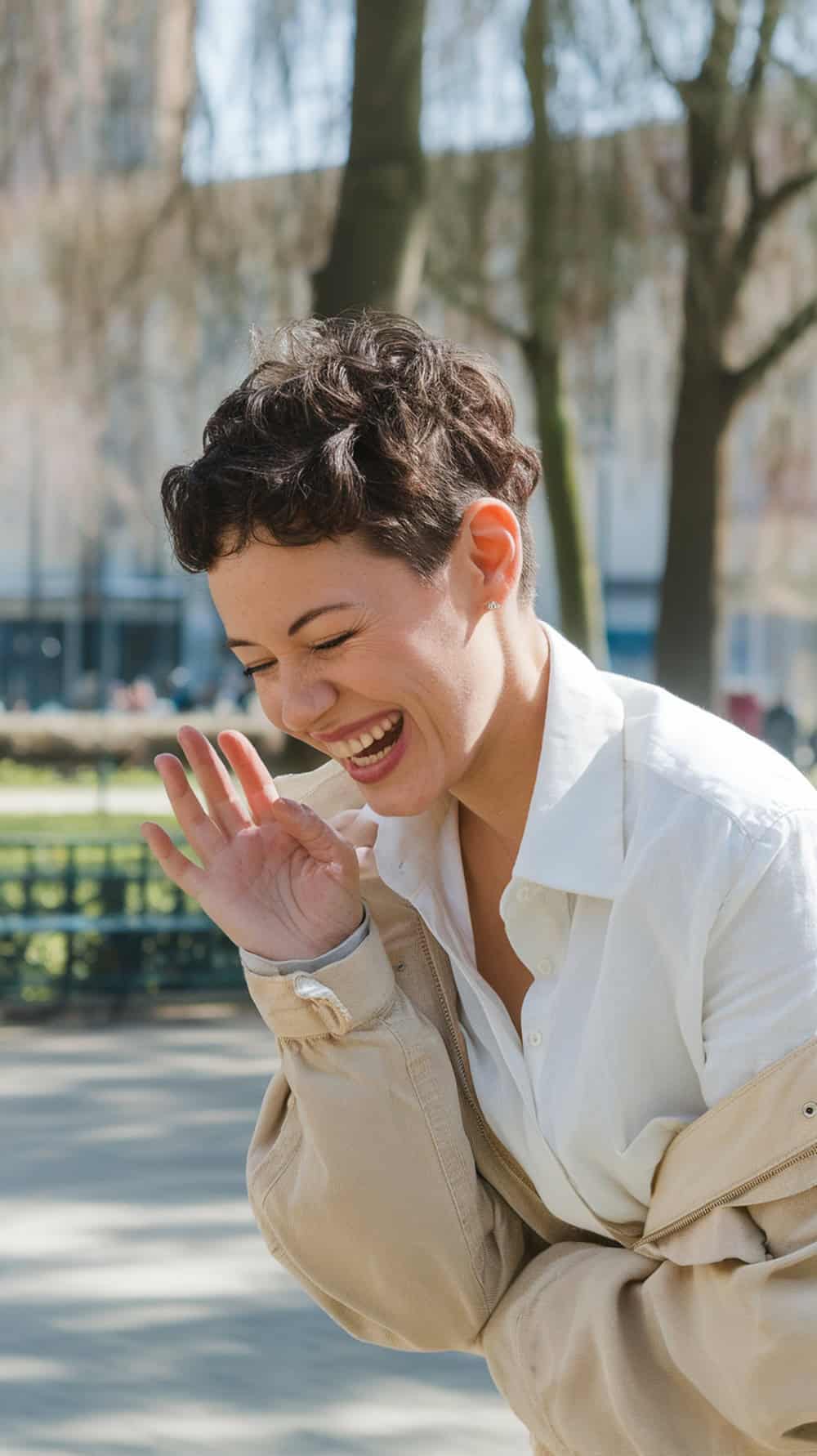A woman with a curly pixie haircut laughing joyfully in a park.