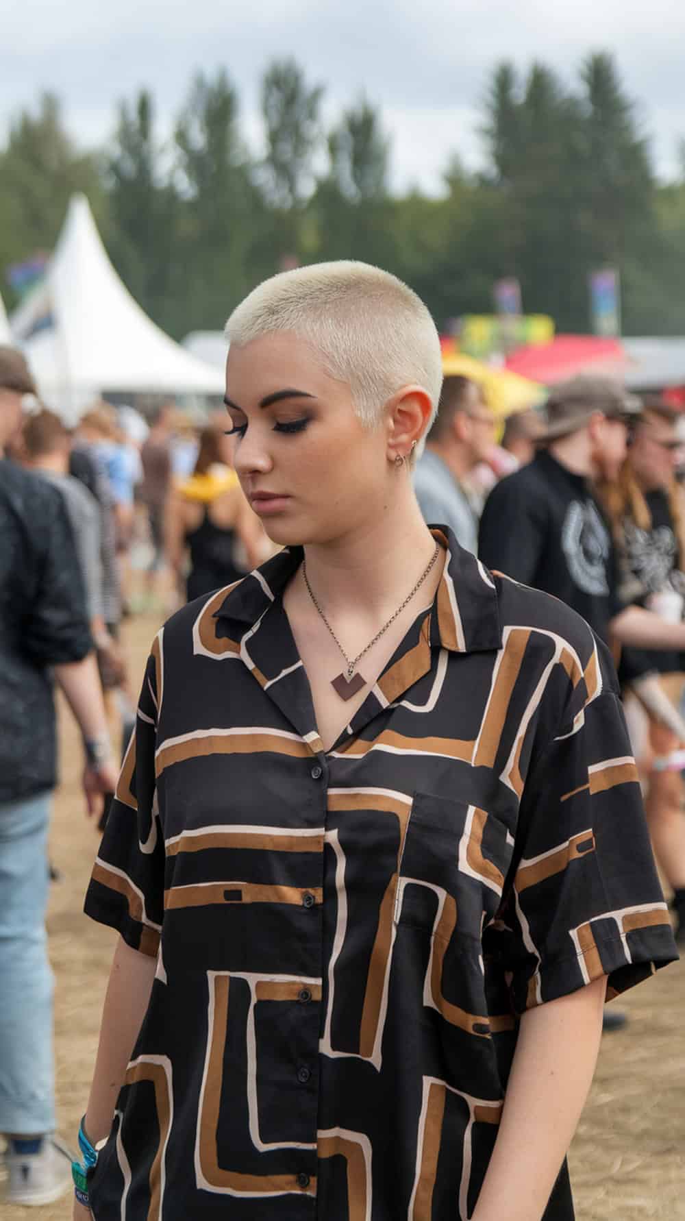 A young woman with a pixie haircut and shaved sides, wearing a patterned shirt at a festival.