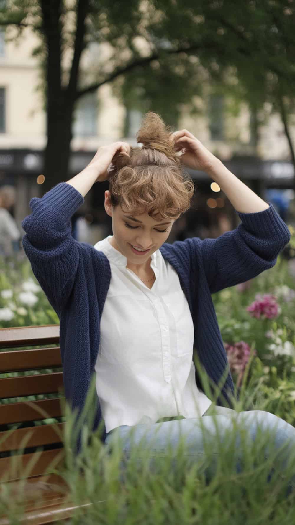 A woman with a curly pixie haircut sitting on a bench in a park, adjusting her hair.
