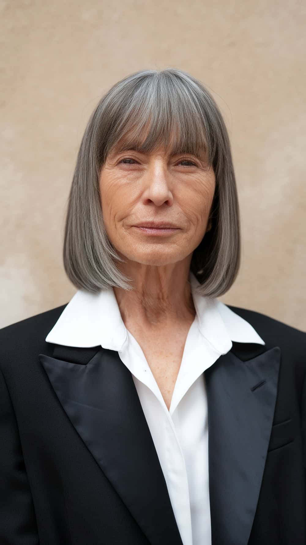 A woman with sleek straight gray hair and bangs, dressed in a black blazer and white shirt, looking confidently at the camera.