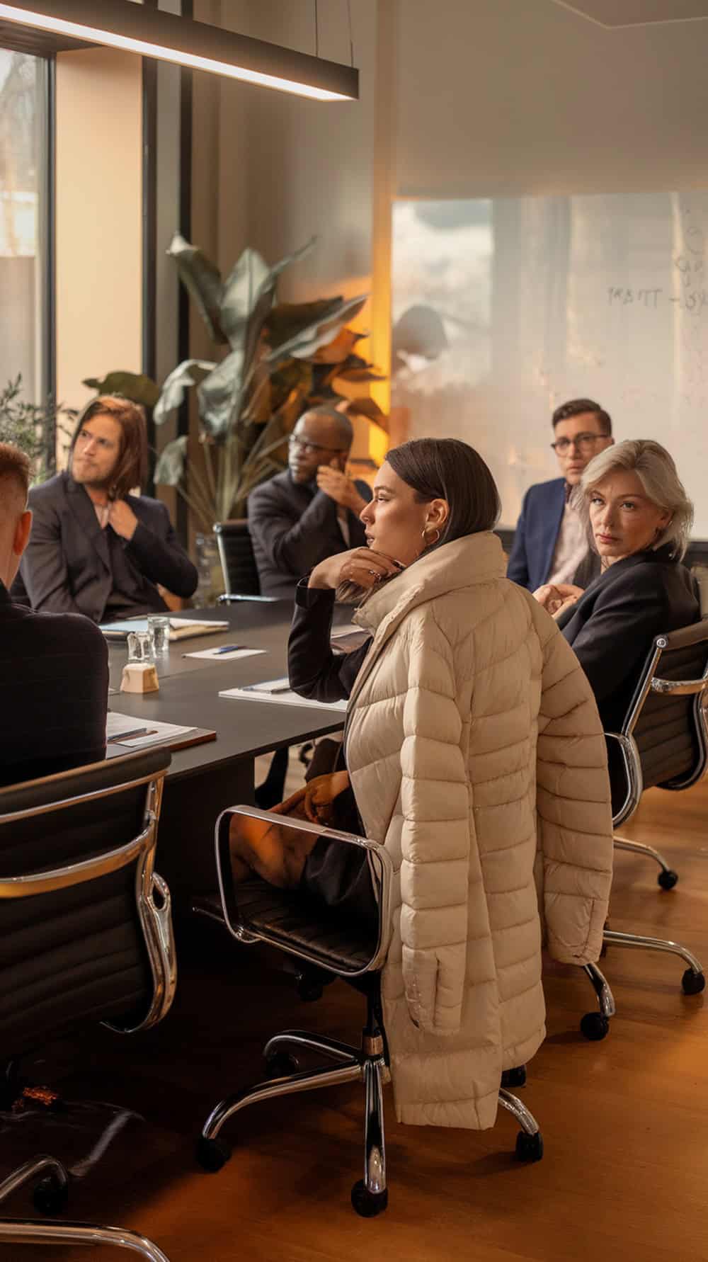 A group of professionals in a meeting, with one woman wearing a stylish puffer coat.