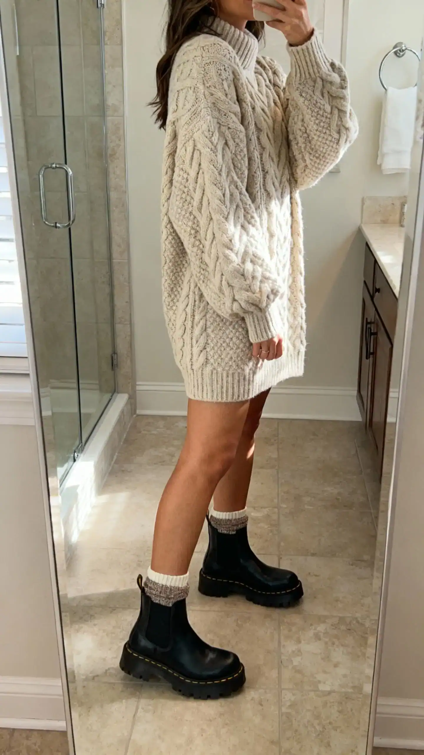 iPhone mirror selfie of a woman wearing a chunky cable-knit sweater dress with lug-sole Chelsea boots and cozy crew socks slightly peeking, face not visible, side angle in a bathroom full-length mirror, soft indoor morning light, iPhone photo quality.