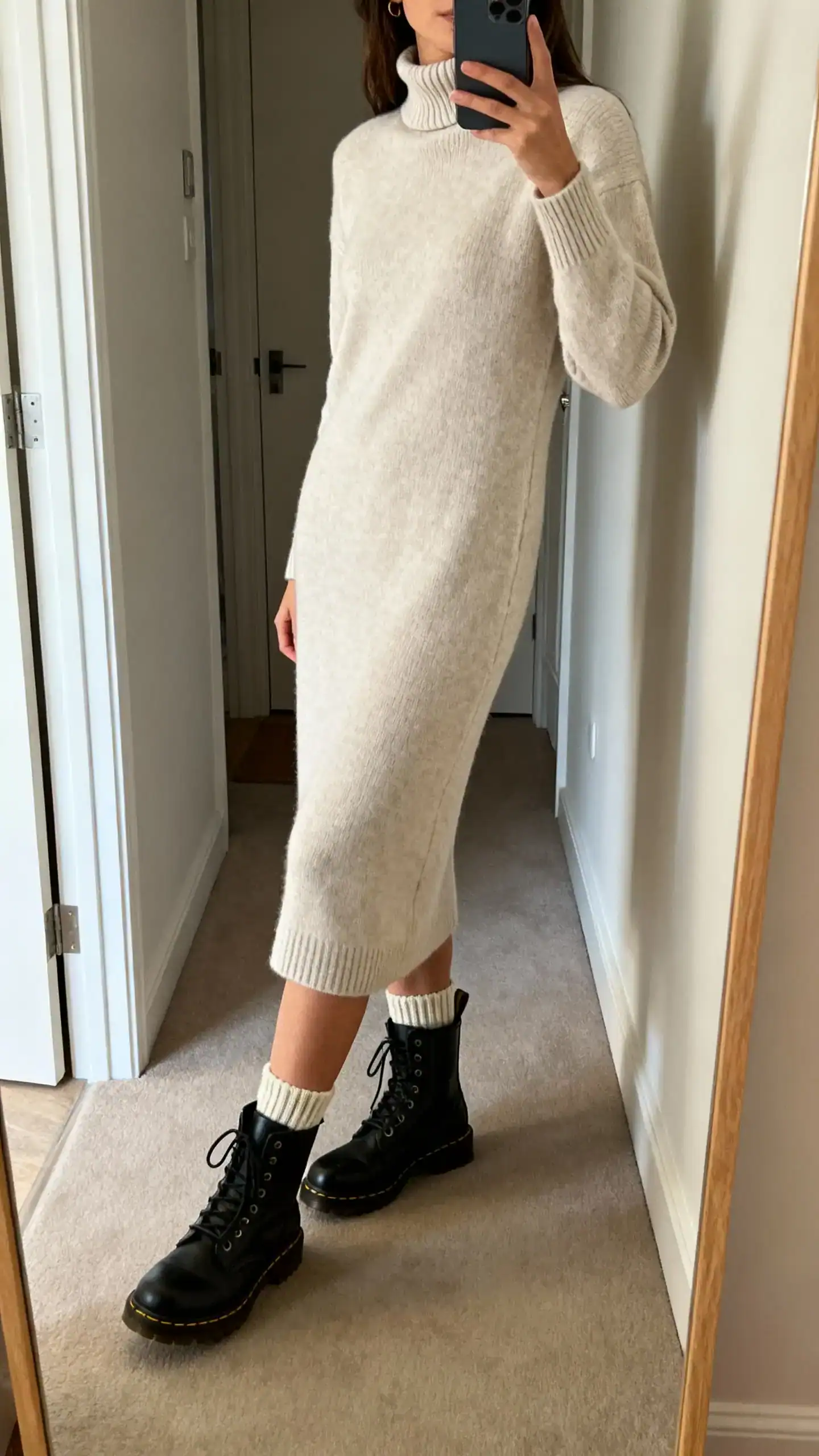 iPhone side-angle mirror selfie of a woman wearing a minimal mock-neck midi sweater dress with combat boots and ribbed ankle socks, face not visible, full-length mirror in entryway, soft indoor lighting, iPhone photo quality.