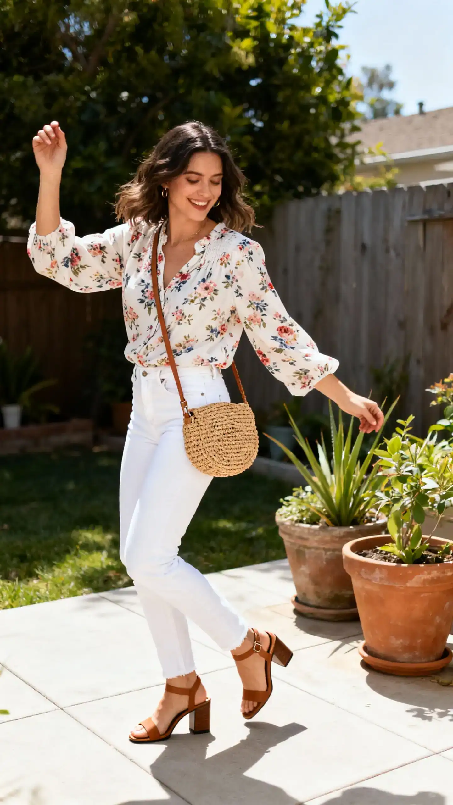 A woman wearing white jeans with a floral blouse, block-heel sandals, and a woven crossbody bag, twirling on a backyard patio near potted plants, casual iPhone photo style, sunny day, outdoor setting.