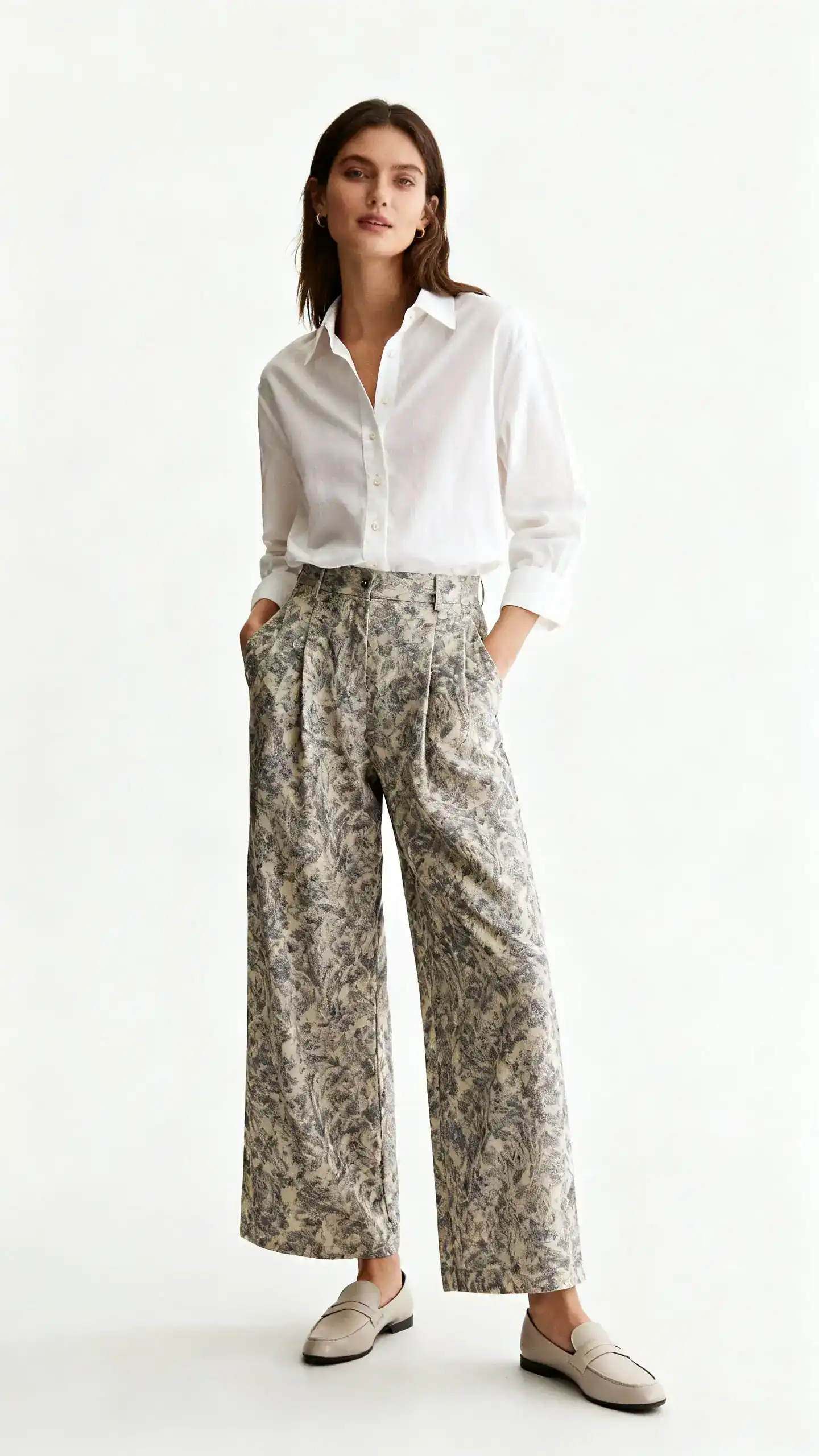 Editorial style studio photo of a woman wearing patterned wide-leg pants with a crisp button-down shirt and simple loafers, white background, soft studio lighting, hands-in-pockets relaxed pose, clean composition, magazine-quality.