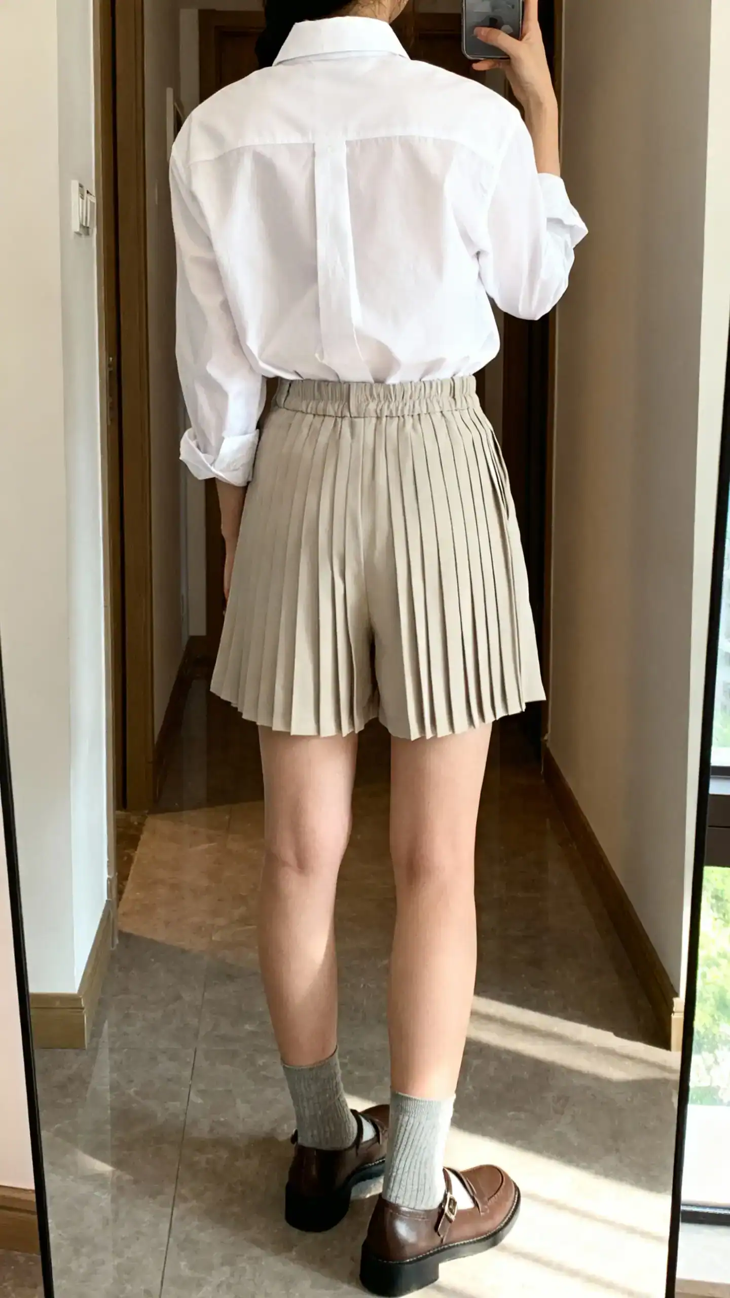 iPhone back-view mirror selfie of a woman wearing a men’s-style white Oxford shirt tucked into pleated shorts with Mary Janes and ankle socks, face not visible, hallway mirror, morning natural light, iPhone photo quality.
