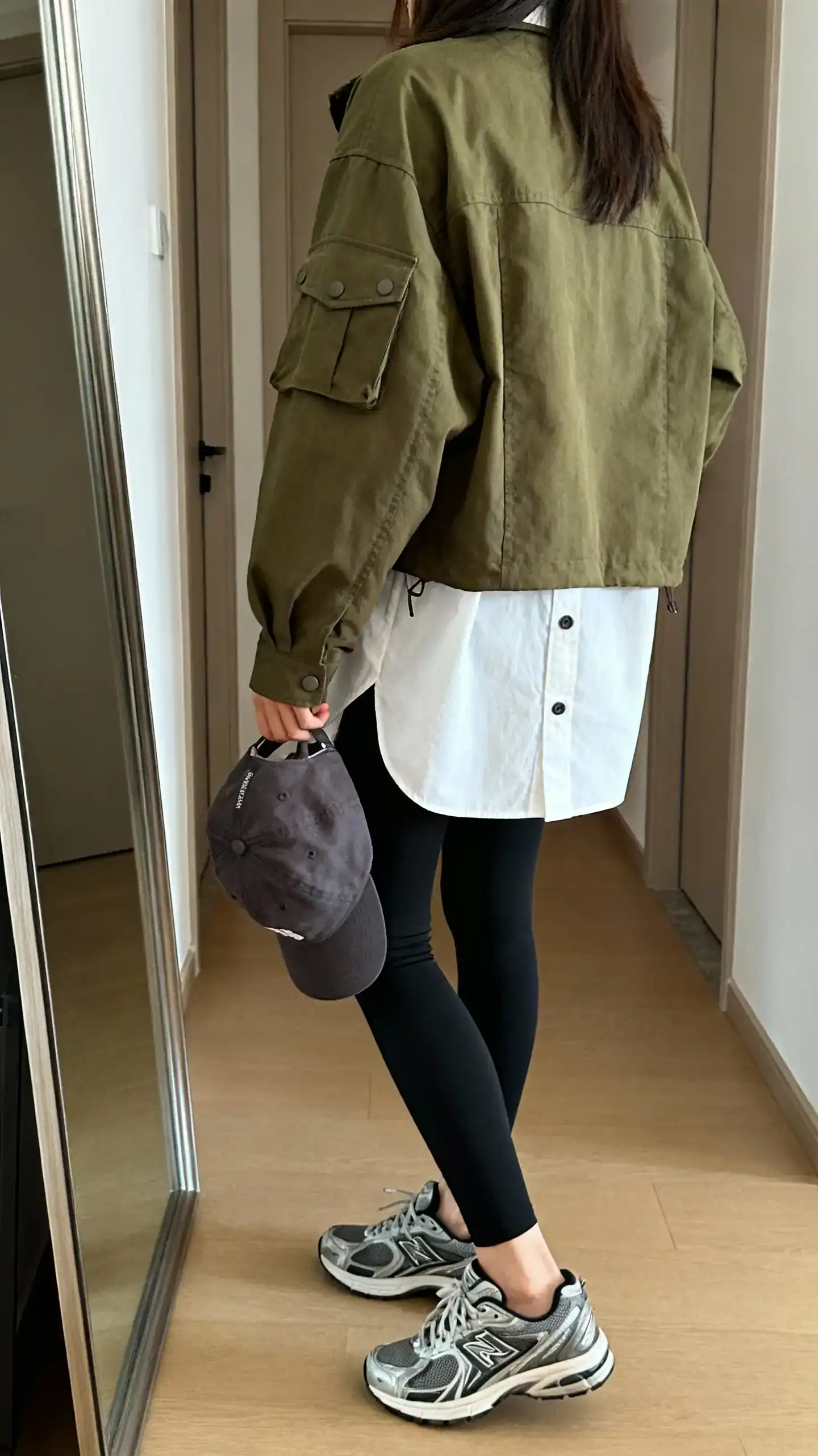 iPhone back view of a woman wearing an olive utility jacket over a white button-up shirt with black leggings, sporty sneakers, baseball cap in hand, face not visible, hallway full-length mirror, natural daylight, iPhone photo quality.