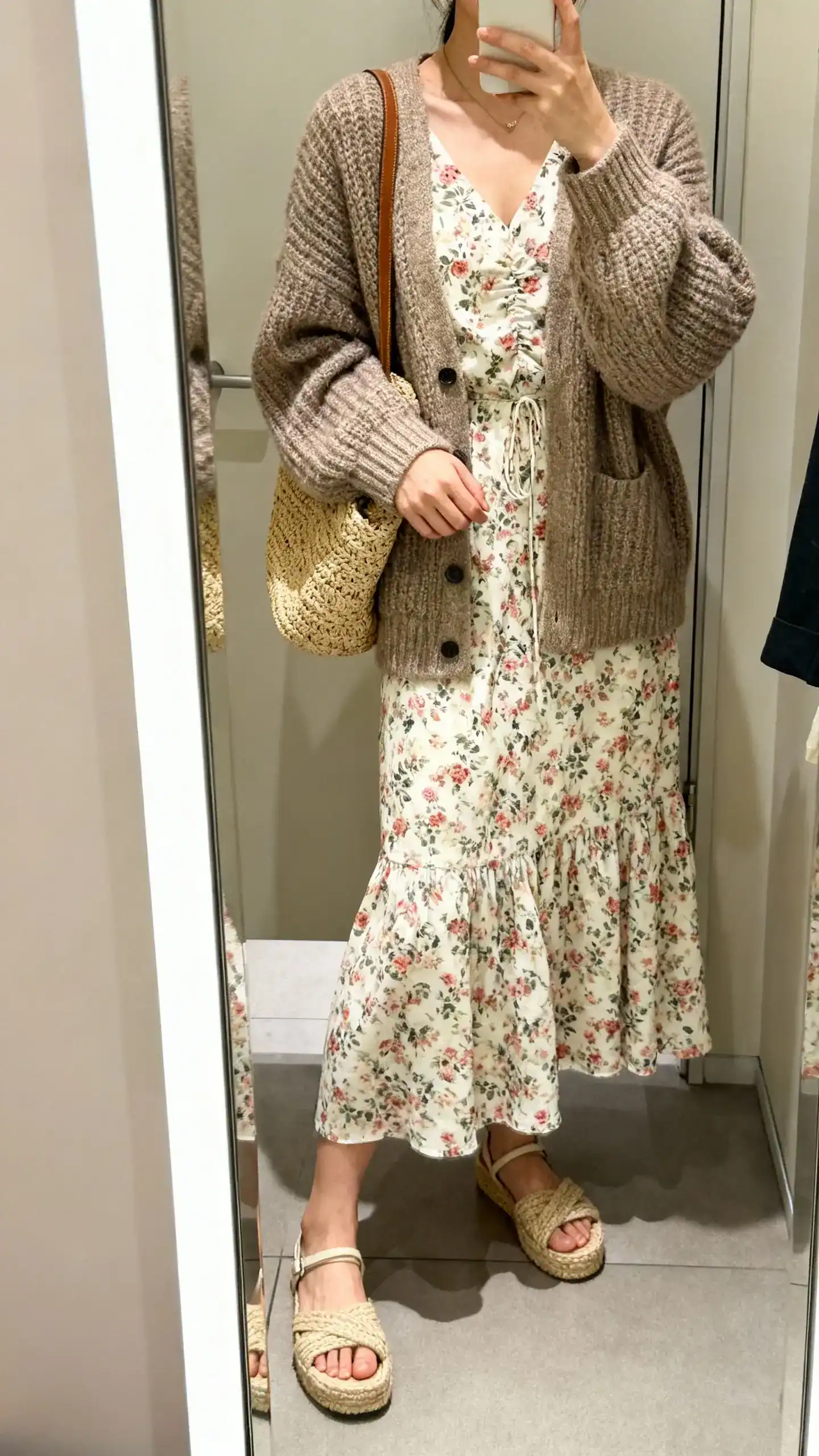 iPhone mirror selfie of a woman in a floral midi dress layered with a chunky cardigan, espadrille sandals, woven tote, face not visible, cropped at neck, dressing room mirror, soft indoor lighting, iPhone photo quality.