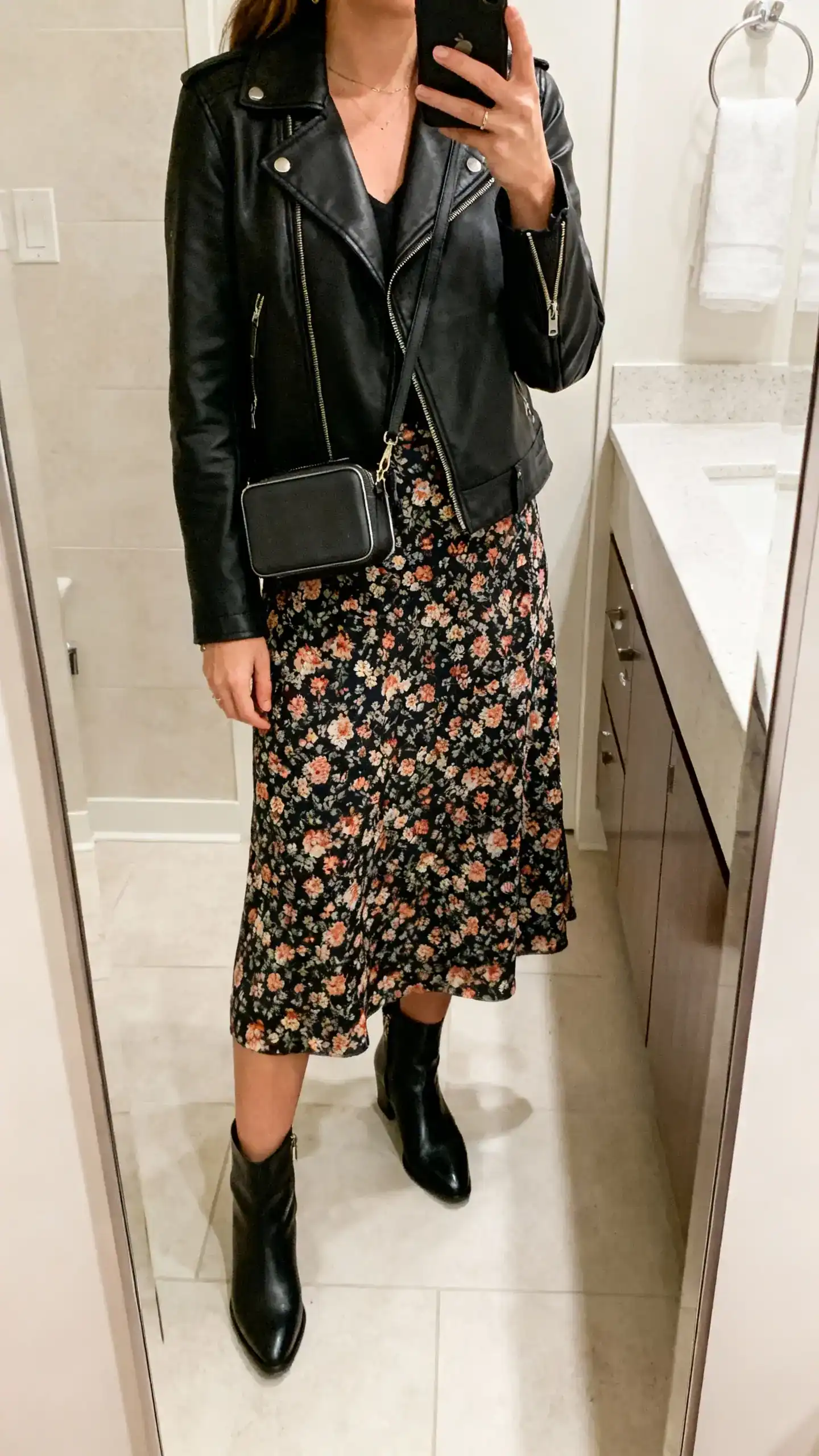 iPhone mirror selfie of a woman in a floral midi skirt with a black leather jacket, ankle boots, and a compact crossbody, face not visible, cropped at neck, bathroom mirror, soft indoor lighting, iPhone photo quality.
