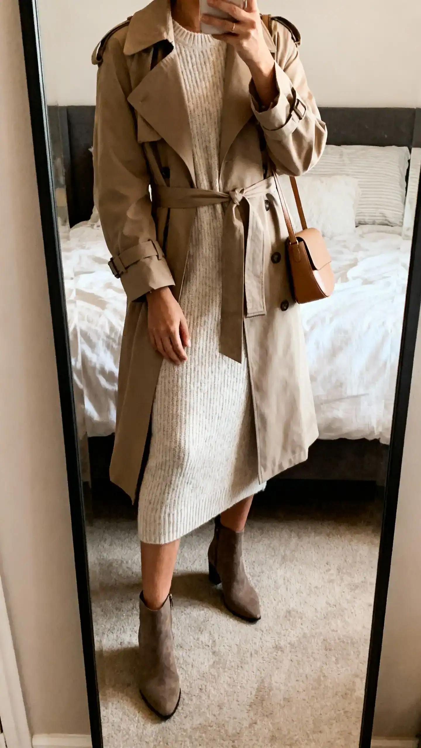 iPhone mirror selfie of a woman in a soft knit dress under a belted trench with ankle boots and a minimal leather bag, face not visible, bedroom full-length mirror, warm indoor lighting, iPhone photo quality.