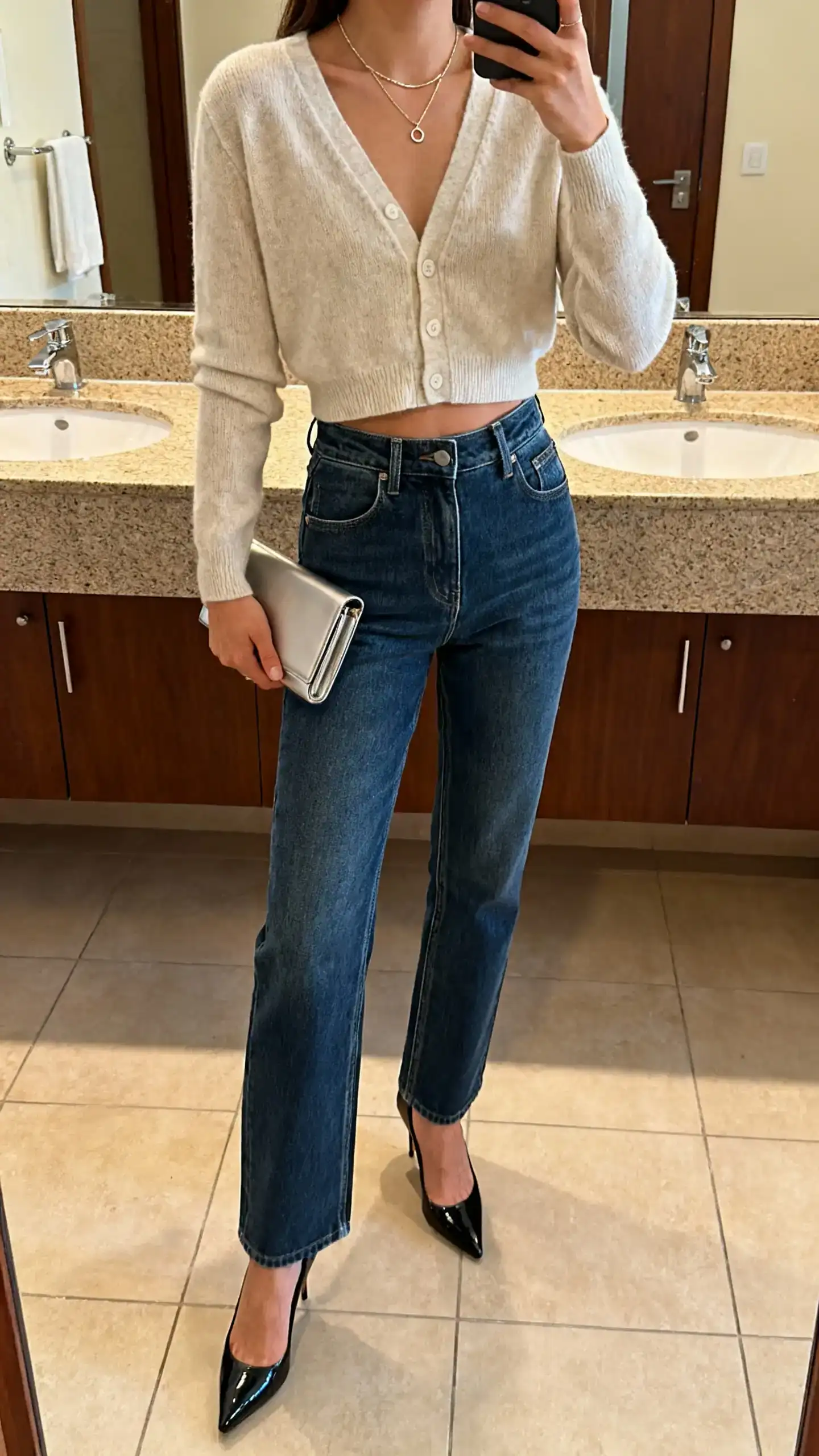 iPhone mirror selfie of a woman styling a cardigan as a top tucked into high-rise straight jeans with sleek heels, dainty necklace, slim clutch, face not visible, cropped at shoulders, bathroom mirror, warm indoor lighting, iPhone photo quality.