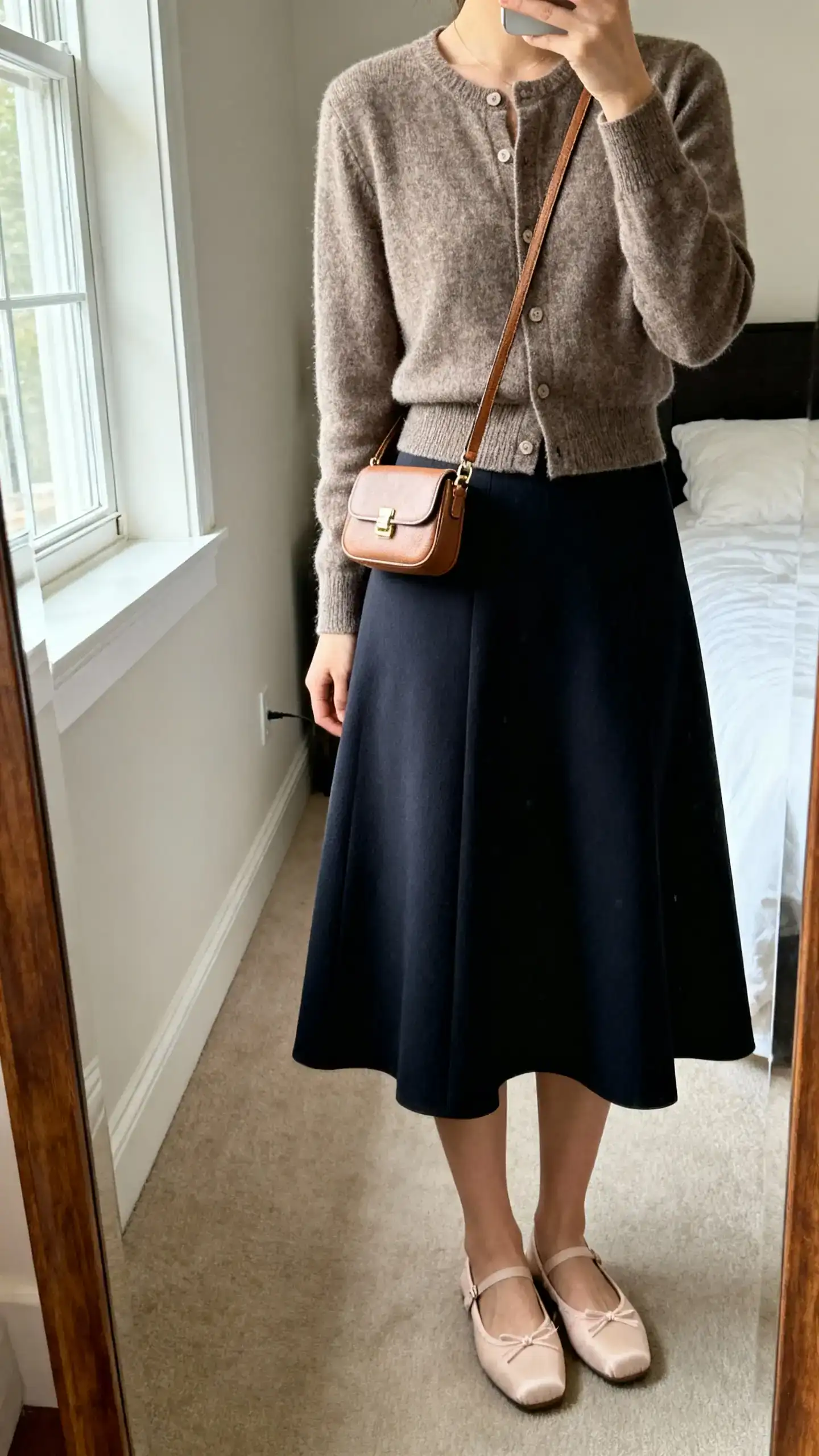 iPhone mirror selfie of a woman wearing a cardigan as a top tucked into an A-line skirt with ballet flats and a petite crossbody, face not visible, cropped at neck, bedroom mirror, natural window light, iPhone photo quality.