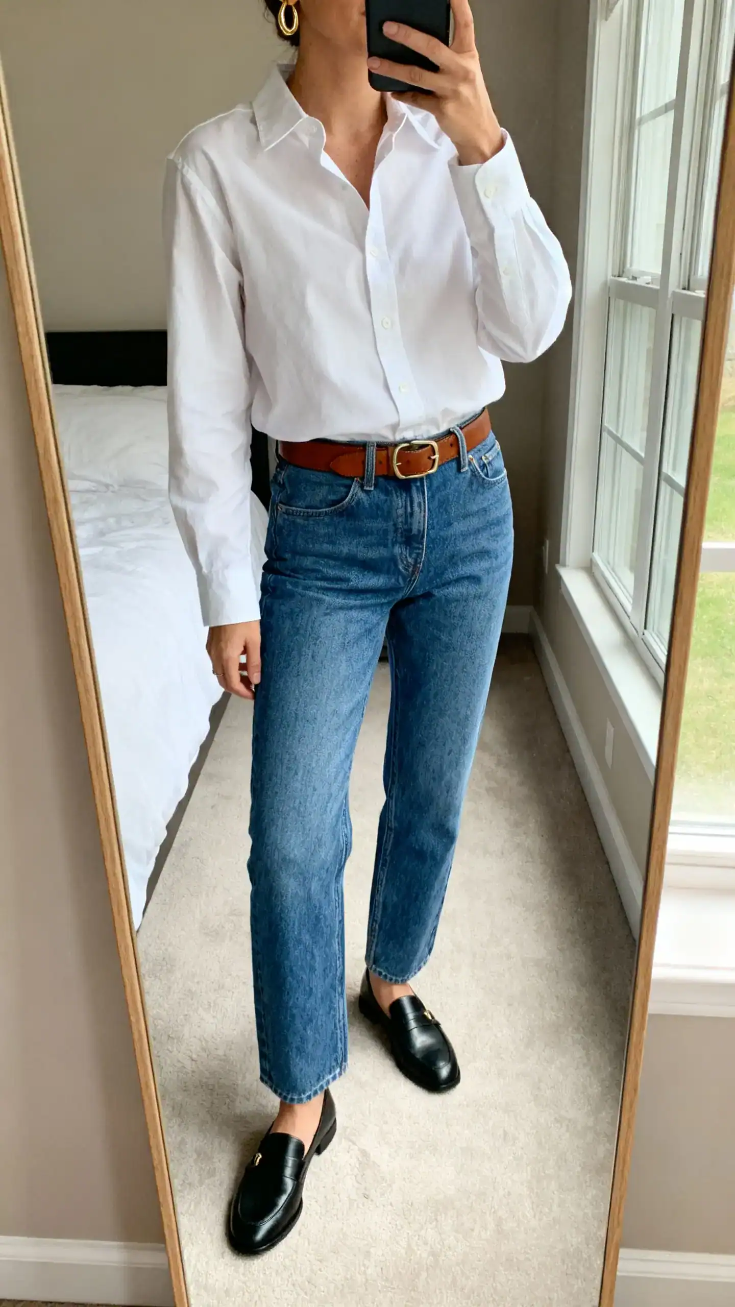 iPhone mirror selfie of a woman wearing a crisp white button-up shirt tucked into classic blue straight-leg denim, brown leather belt, minimal gold hoops, black loafers, face not visible, cropped at neck, bedroom mirror with natural window light, iPhone photo quality.