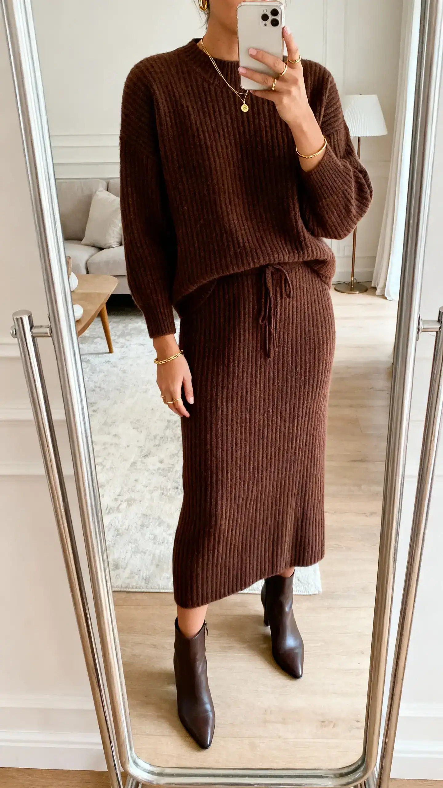 iPhone mirror selfie of a woman wearing a monochrome knit set (matching ribbed sweater and midi skirt) in chocolate brown, sleek ankle boots, subtle gold jewelry, face not visible, cropped at shoulders, living room full-length mirror, soft indoor light, iPhone photo quality.