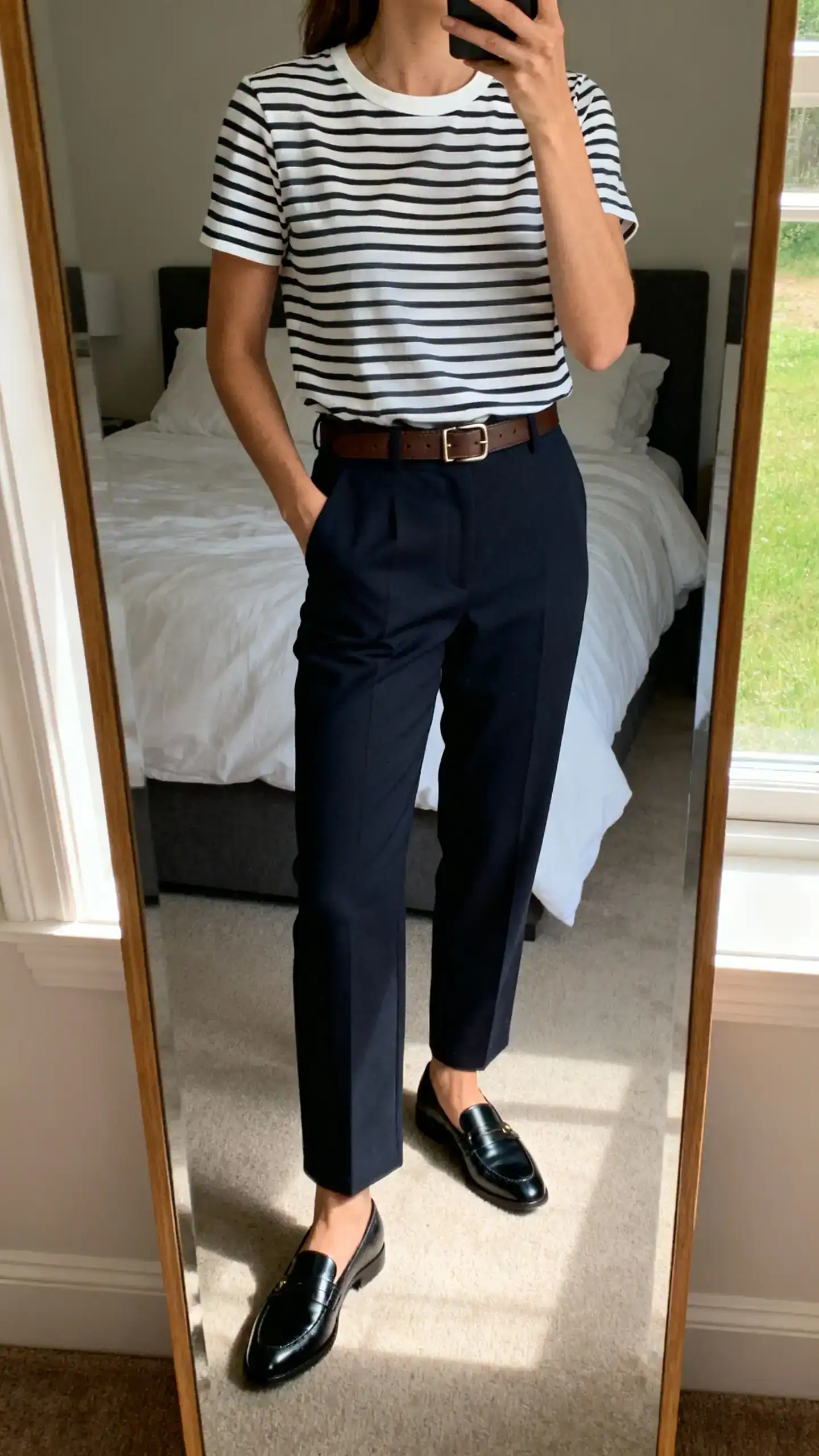 iPhone mirror selfie of a woman wearing a striped Breton tee tucked into tailored trousers with sleek loafers and a simple leather belt, face not visible, cropped at neck, bedroom mirror, morning window light, iPhone photo quality.