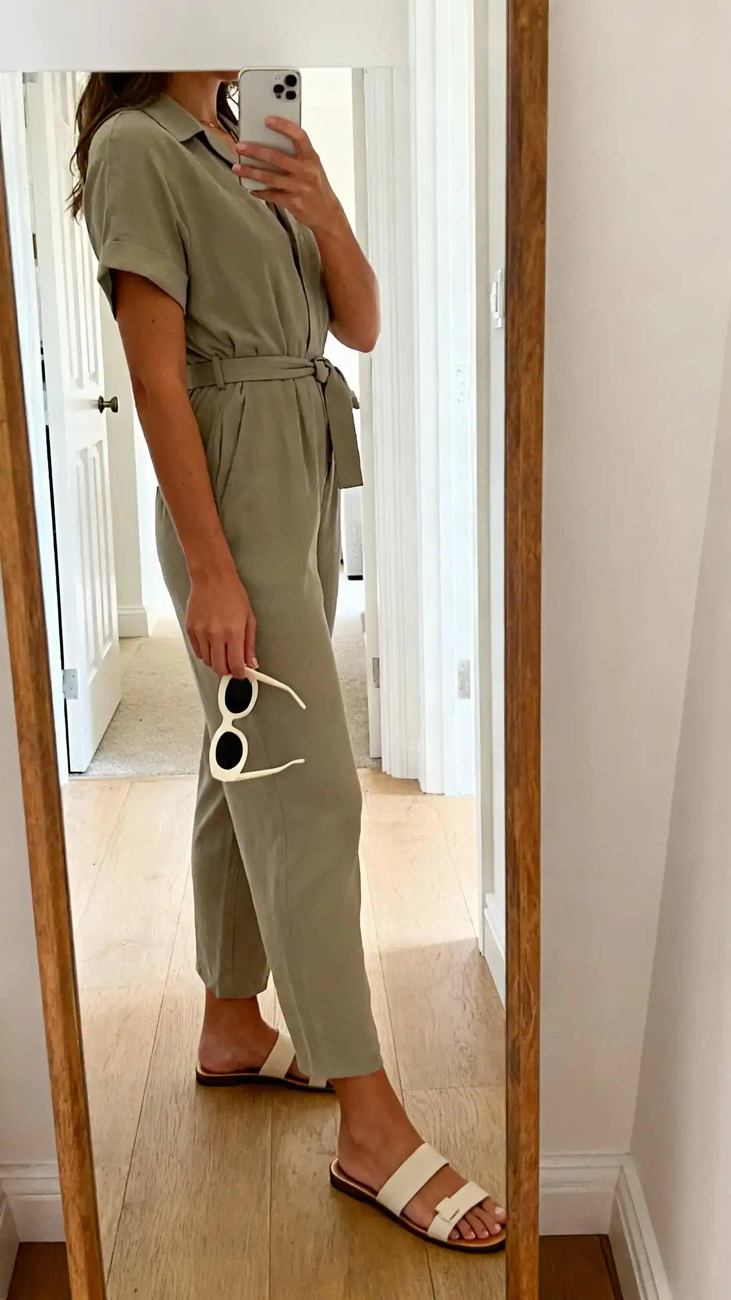 iPhone side-angle mirror selfie of a woman in a minimal jumpsuit cinched with a belt, statement sunglasses in hand, and sleek sandals, face not visible, entryway mirror, natural daylight, iPhone photo quality.