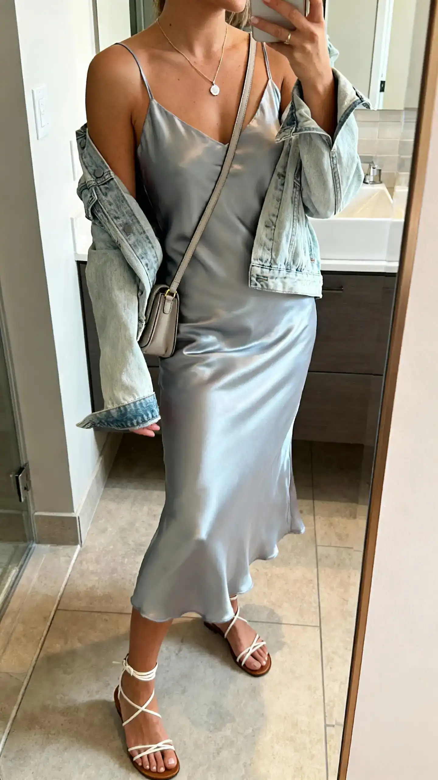 iPhone side-angle mirror selfie of a woman in a satin slip dress with a light-wash denim jacket draped over shoulders, delicate pendant necklace, ankle-strap sandals, small crossbody bag, face not visible, bathroom full-length mirror, morning light, iPhone photo quality.