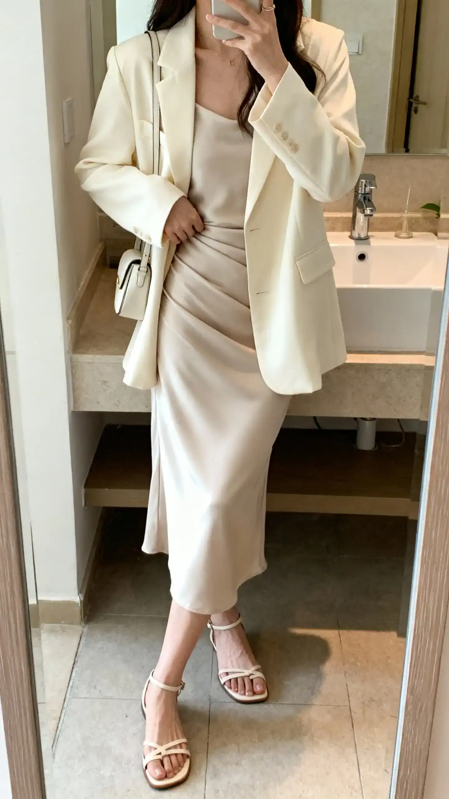 iPhone side-angle mirror selfie of a woman wearing a cream blazer layered over a bias-cut slip dress with delicate sandals and a small shoulder bag, face not visible, bathroom mirror, soft indoor lighting, iPhone photo quality.