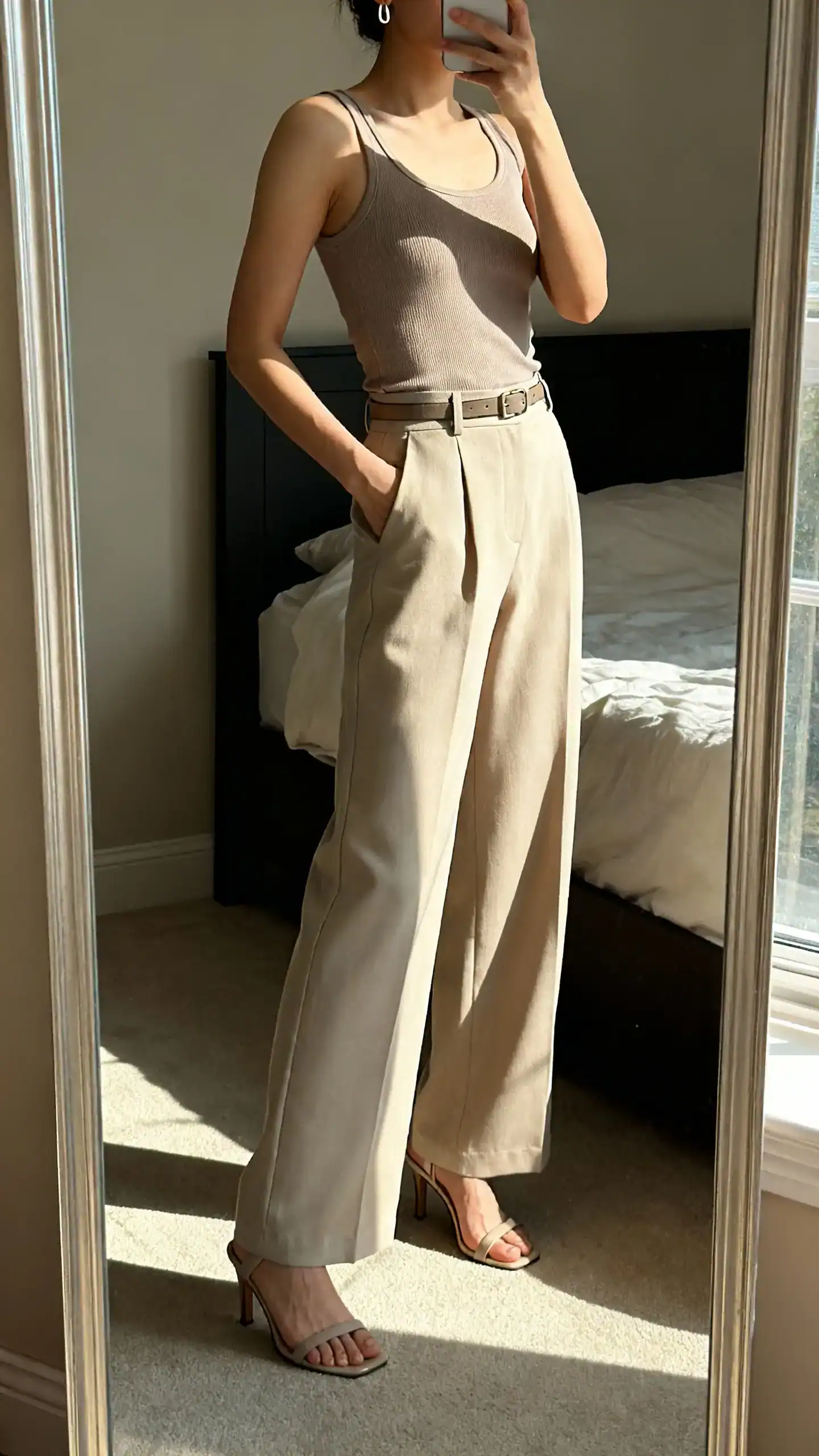 iPhone side-angle mirror selfie of a woman wearing a fitted tank top with high-waist wide-leg trousers, slim belt, heeled sandals, minimalist earrings, face not visible, cropped at shoulders, bedroom mirror, morning window light, iPhone photo quality.