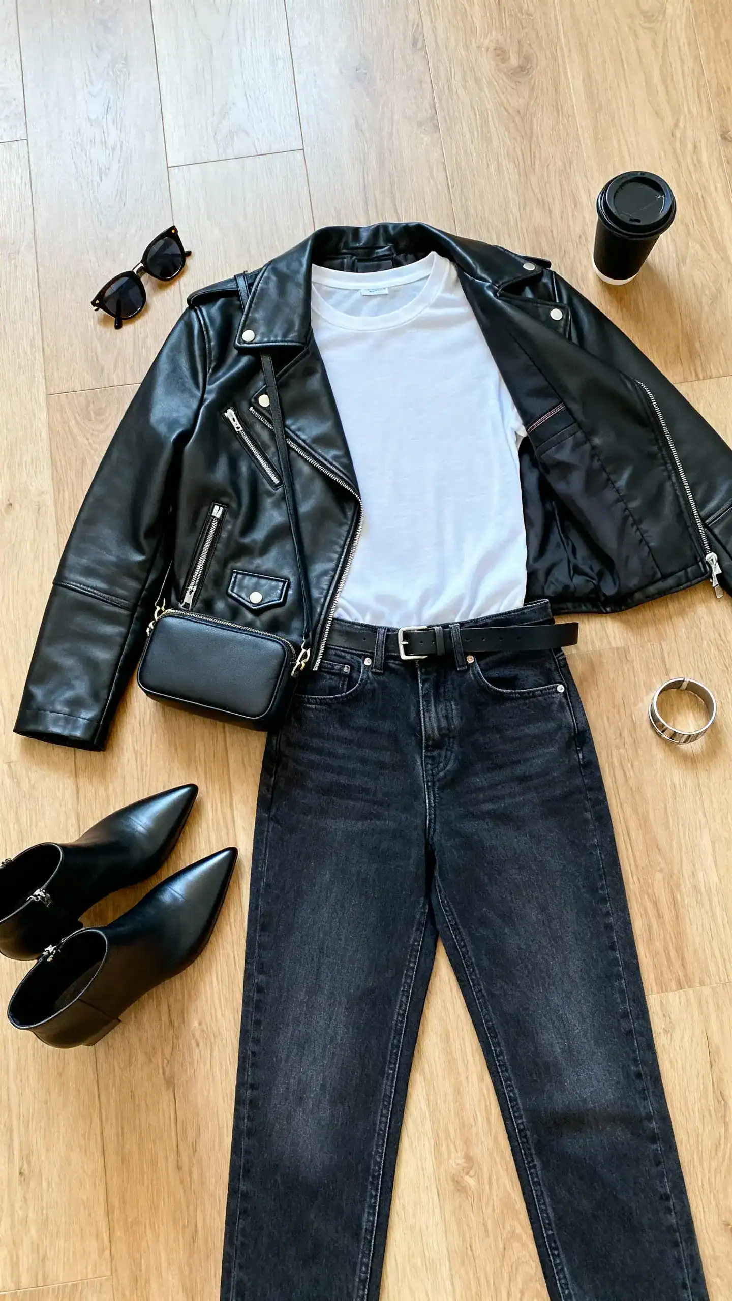 Top-down flat lay of a black lightweight leather moto jacket, high-rise straight jeans, fitted crewneck tee, pointed ankle boots, compact crossbody, bold sunglasses, and a minimal belt on a wooden floor; include a black coffee and a metal cuff bracelet, bright natural light, no person visible, organized composition, iPhone photo aesthetic.
