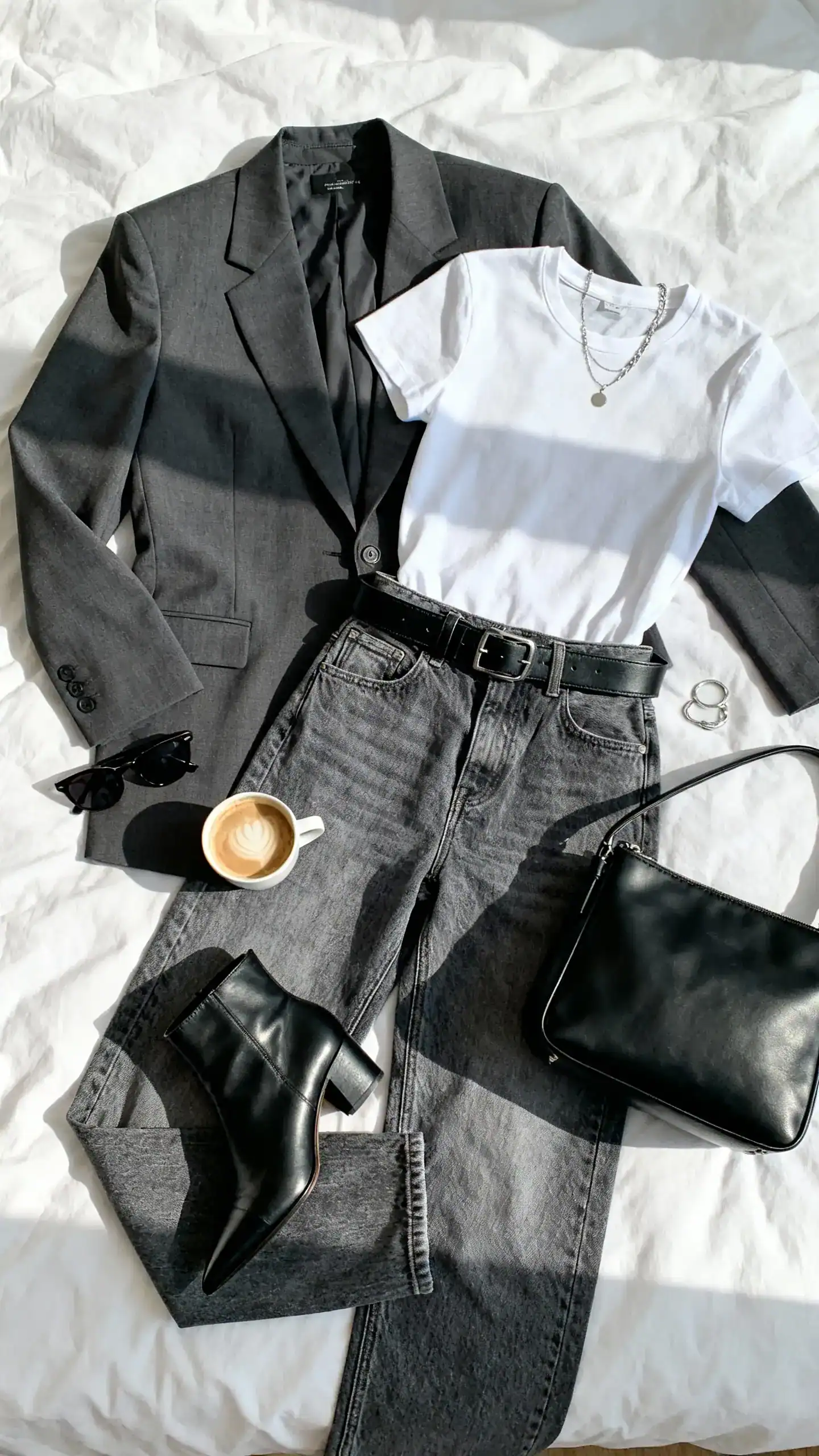 Top-down flat lay of a sharp charcoal power blazer, relaxed mid-wash straight-leg denim, white crewneck tee, black leather belt, pointed black ankle boots, sleek black tote, and minimalist silver jewelry on a white bed; add sunglasses and a latte, bright natural light, no person visible, organized composition, iPhone photo aesthetic.