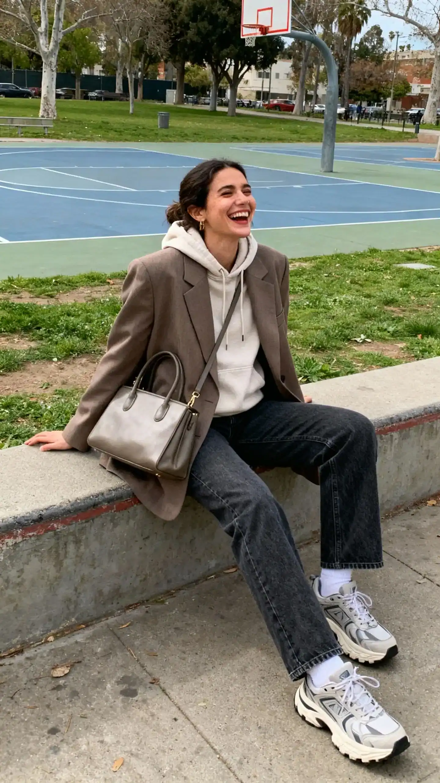 A woman wearing an oversized blazer layered over a hoodie with straight jeans, chunky sneakers, and a structured tote, laughing while sitting on a low wall near a park basketball court, casual iPhone photo style, natural daylight, outdoor setting.