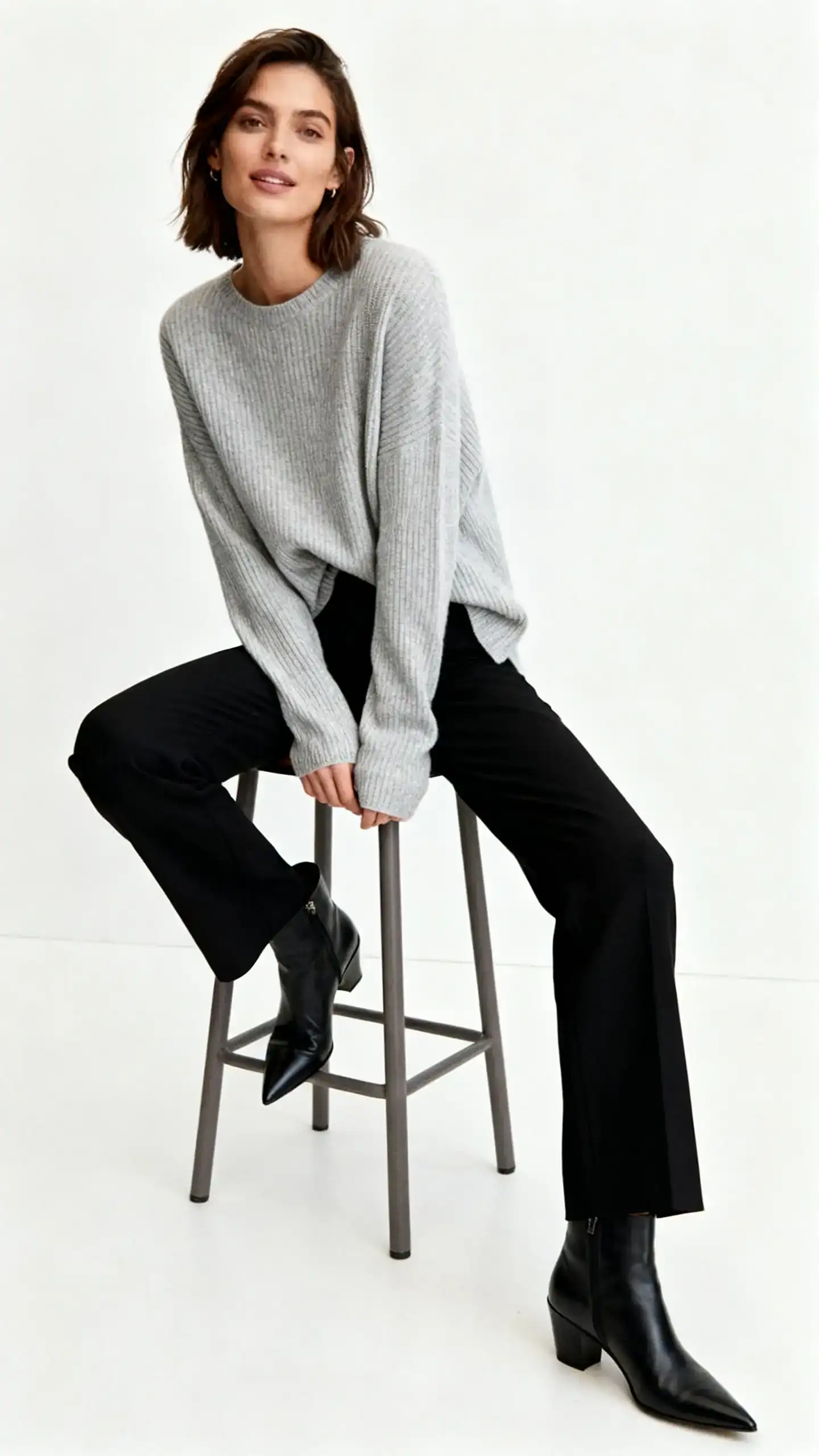 Editorial studio photo of a woman in black trousers paired with a soft light-gray knit sweater (fine gauge, slightly draped) and pointed black ankle boots, white background, studio lighting, natural seated pose on a simple stool, clean composition, desk-to-dinner vibe.